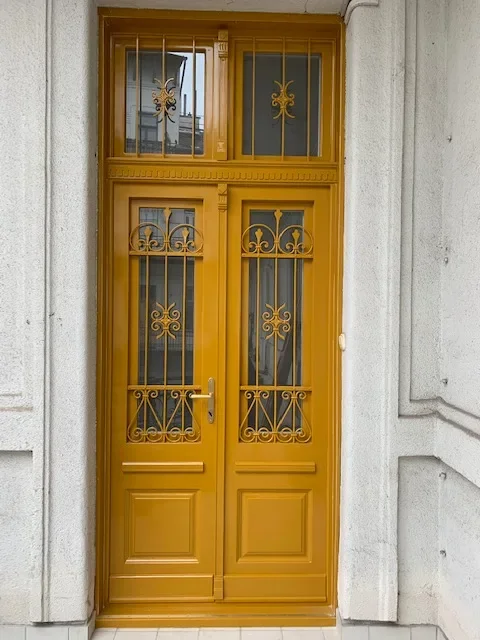 Körfolyosós társasházi bejárati ajtó igazi kovácsoltvas ráccsal, az ajtóban nyitható ablakokkal. Felül nyitható ablakokkal, hőszigetelt üveggel, dupla gumiszigeteléssel, kívül sárgára fényezve, kompletten, beépítéssel együtt.