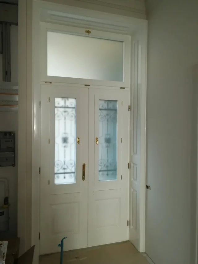 Körfolyosós társasházi bejárati ajtó igazi kovácsoltvas ráccsal, az ajtóban nyitható ablakokkal. Felül bukó kivitelű, kitámasztóval, hőszigetelt üveggel, dupla gumiszigeteléssel, réz kilincsekkel, belül fehérre fényezve, kompletten, beépítéssel együtt. Kazettás beltéri ajtó monarchia rézkilincsekkel, fehérre fényezve.